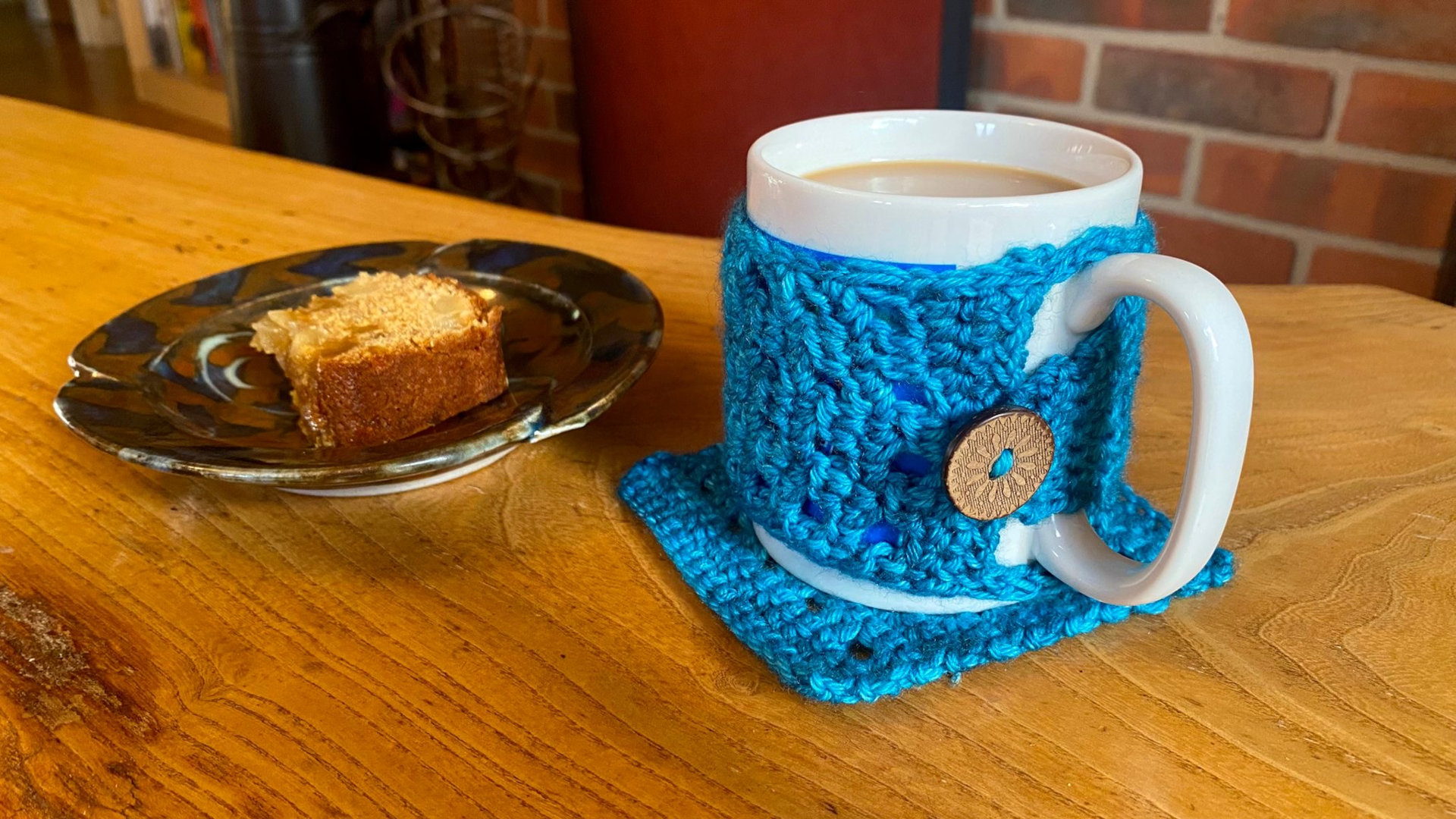 A mug with a crocheted cosy, sat on a crocheted coaster. There is a slice of cake on a plate in the background.