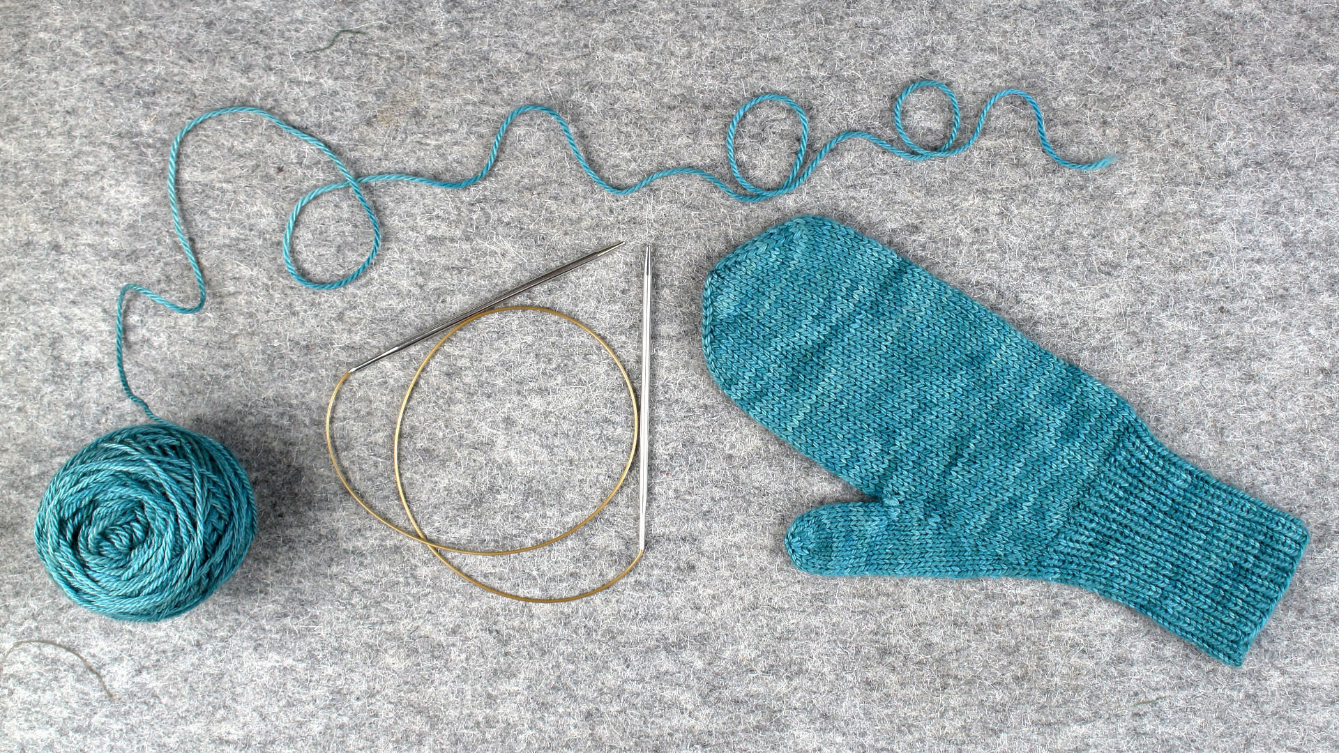 A ball of yarn, a circular needle, and a hand knitted mitten